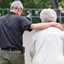 Elderly man standing next to elderly woman with arm draped across her shoulders.