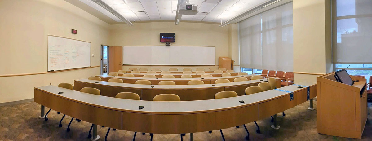 Angled view of a Pharmacy Hall North classroom with curved rows of desks and chairs facing a teaching area with whiteboards and a podium.