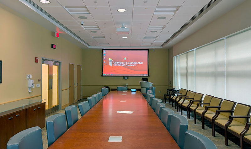 Dean’s Board Room with a long conference table surrounded by chairs, a large projection screen at the front, and additional seating along the right wall.