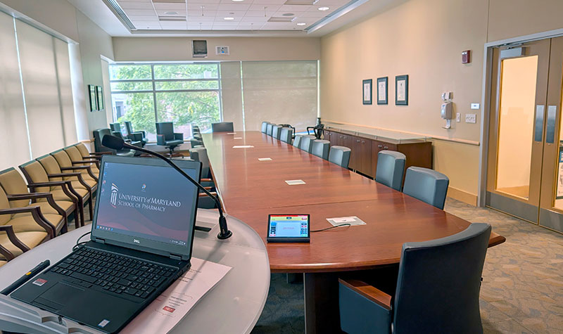 Alternate view of the Dean’s Board Room with a long conference table, rolling chairs, a lectern with laptop and microphone, and large windows at the far end.