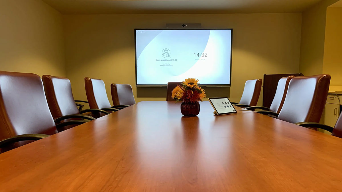 Conference room Saratoga 01‑229 with a long wood conference table, high‑back chairs, a wall‑mounted display screen, and a tablet control panel.